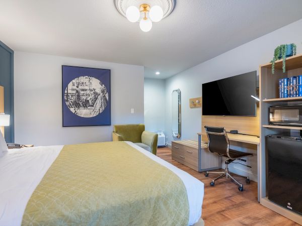 A bright hotel room with a bed, yellow-green throw, wall art, a workspace with desk and TV, and a kitchenette shelf on the right.