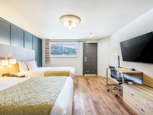 A bright hotel-style room with two beds, a work desk, wall-mounted TV, and a window view, featuring warm lighting and wood flooring.