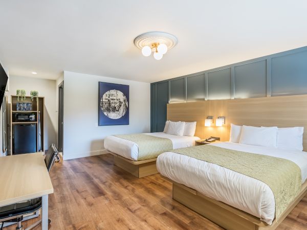 A modern hotel room with two queen beds, a blue padded headboard wall, wooden floors, and a small desk area, feel free to ask for more details.