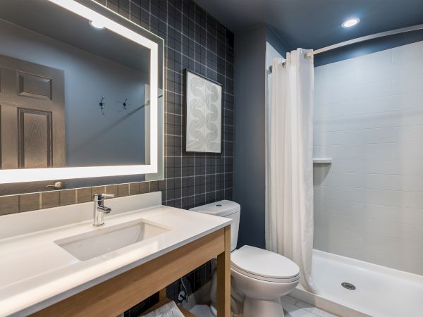 A modern bathroom with a backlit rectangular mirror, dark tile wall, white sink on a wooden vanity, toilet, and a glass-enclosed shower with a white curtain.
