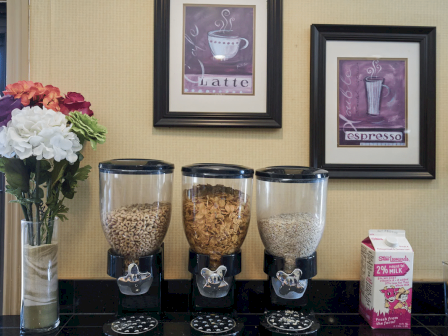 A coffee bar with three cereal dispensers, a vase of flowers, and framed coffee-themed art; a milk carton sits to the right on a dark counter.