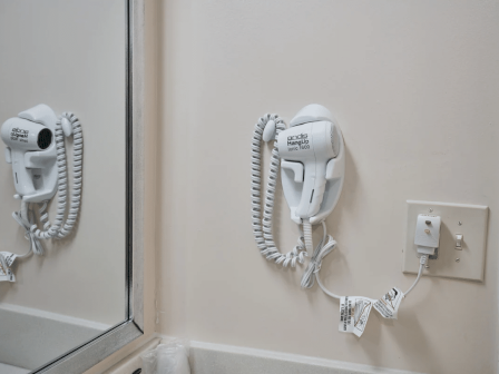 A wall with two mounted hairdryers on coiled cords next to an outlet and a mirror, typical in a hotel bathroom, with light-colored walls.