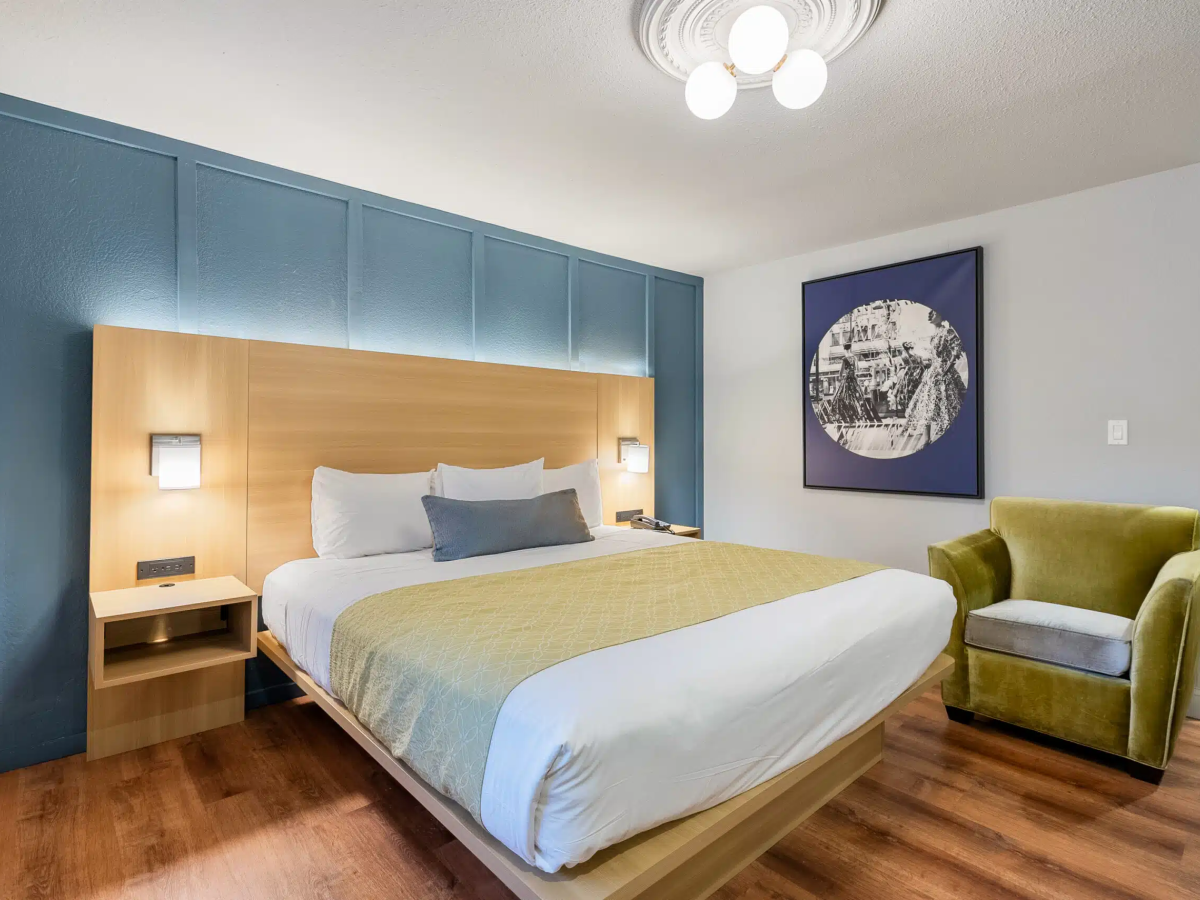 A modern hotel room with a large bed, blue padded wall behind the headboard, wooden nightstands, a mustard-yellow throw, a green armchair, and framed artwork; warm lighting and wooden flooring.