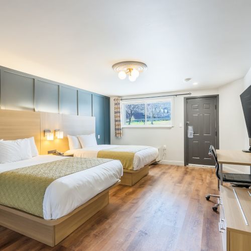 Bright hotel room with two neatly made beds, teal headboards, wooden floors, a desk with TV on the wall, and a window near the door.