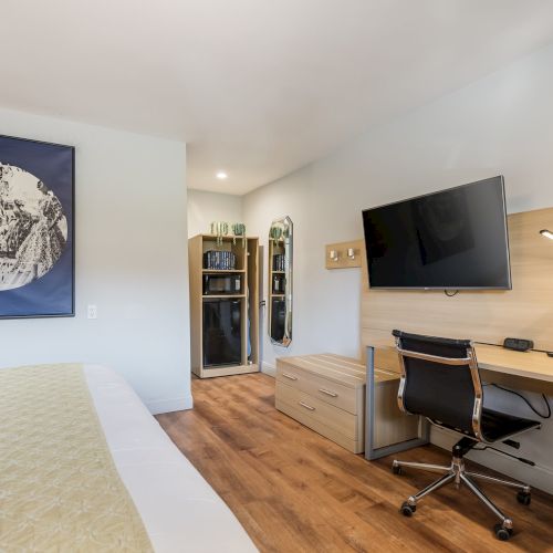A modern hotel-style bedroom with a bed, a wooden desk and chair, a mounted TV, a floor lamp, and a small wardrobe area, plus a blue framed art piece on the wall.