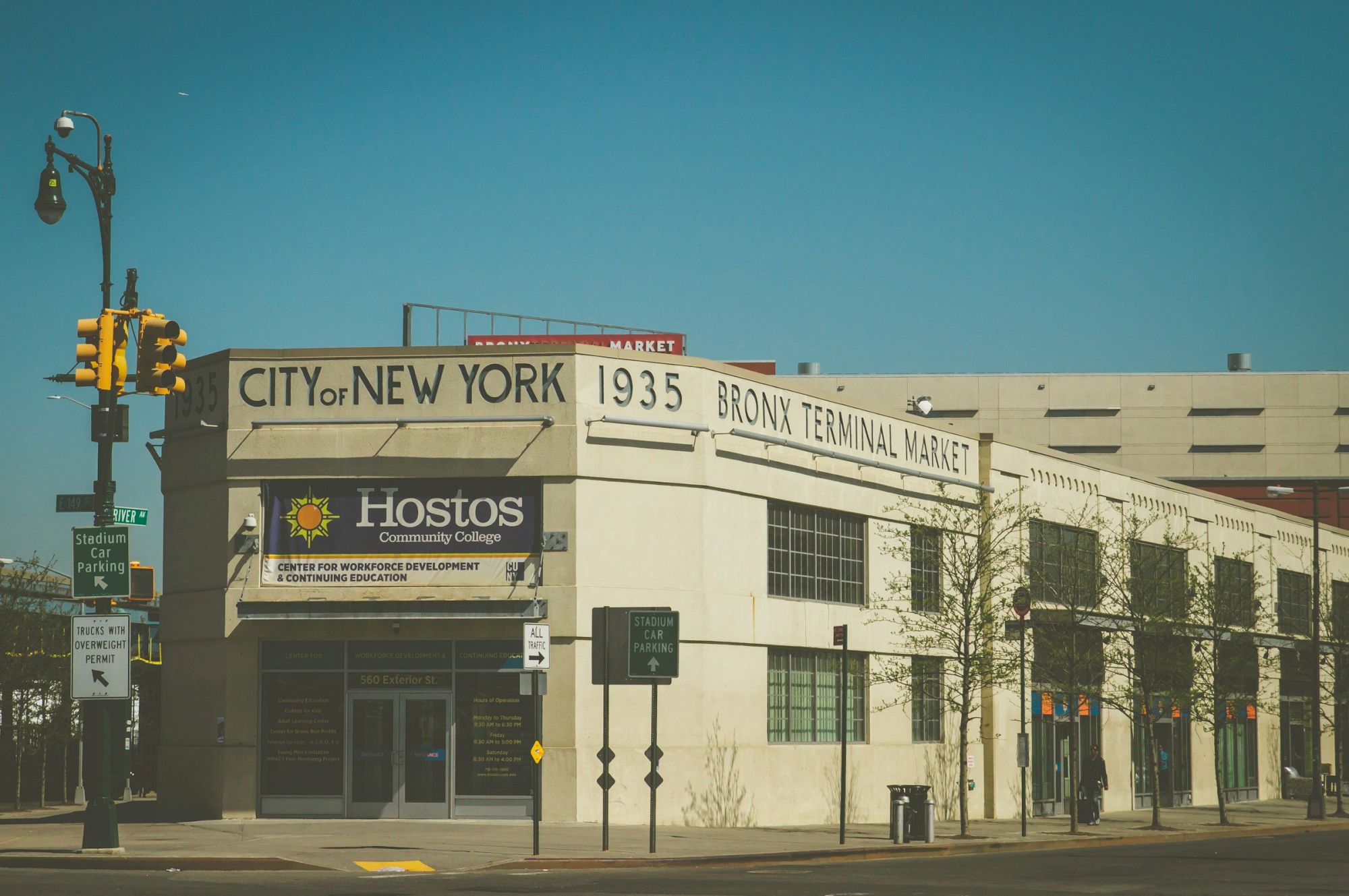 A vintage-style building on a sunny street corner, labeled &ldquo;City of New York 1935 Bronx Terminal Market,&rdquo; with a Hostos sign and palm trees nearby.