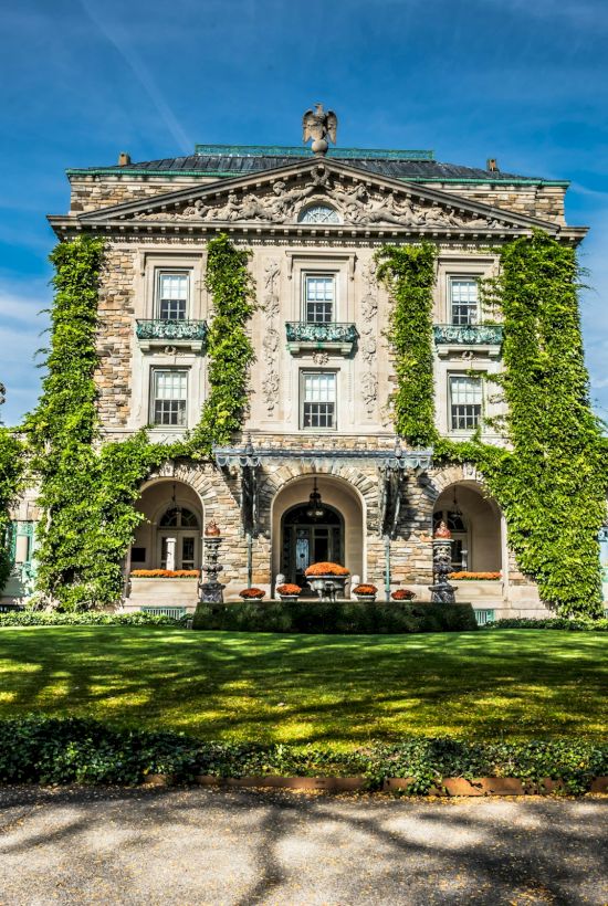 A grand ivy-covered mansion with a stone facade, arched entrance, manicured lawn, and trees framing a sunny blue sky.